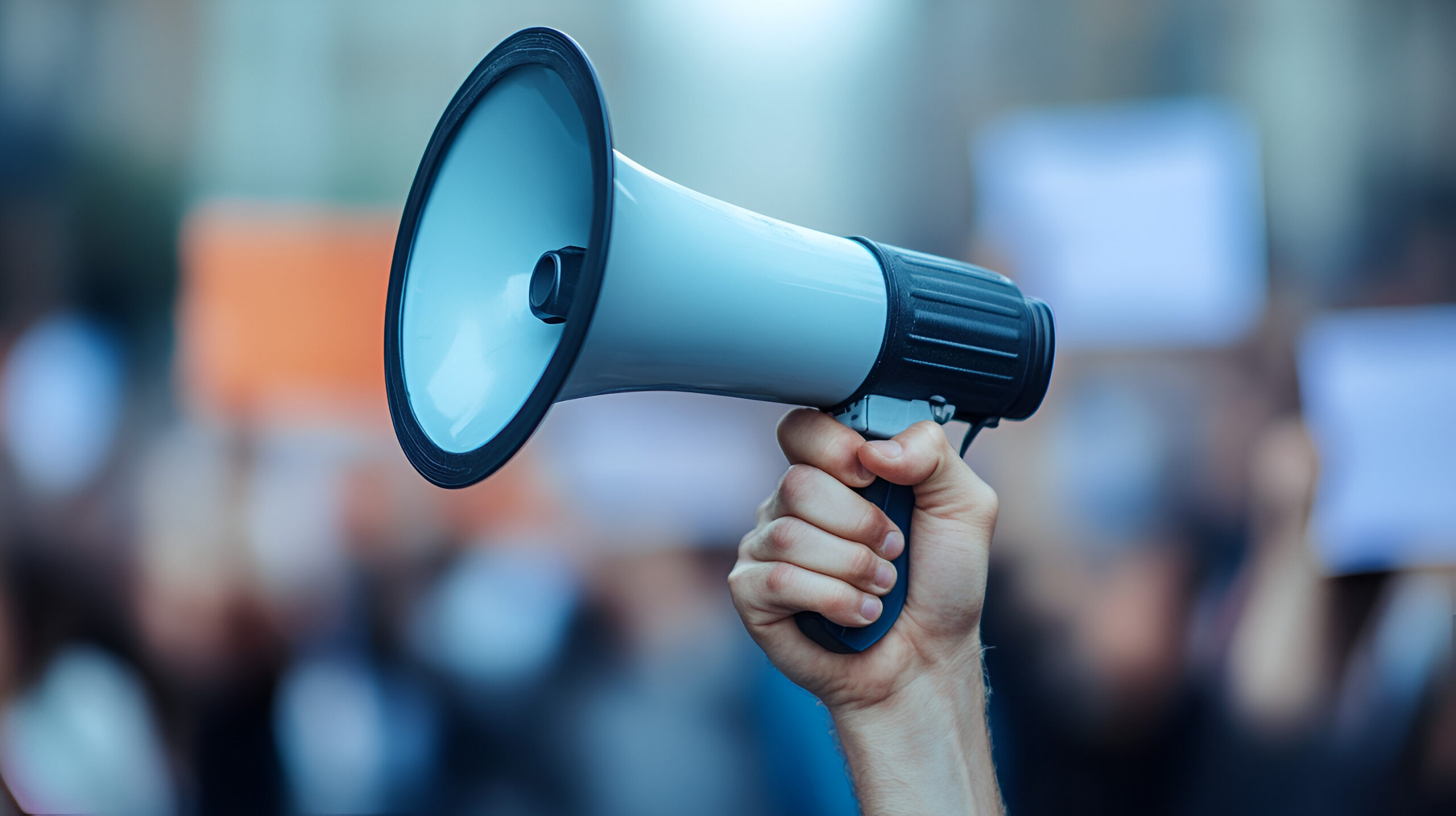 A hand holding up a megaphone against a blurred-out background of a crowd of people with signs.